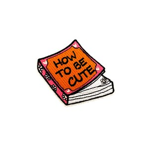May include: Embroidered patch in the shape of a book with the words "HOW TO BE CUTE" in black on an orange cover. The book has a pink border with white flower details and a stack of white pages.