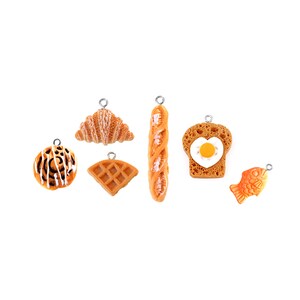 Könnte beinhalten: Ein Set mit sieben Anhänger-Anhängern, die verschiedene Lebensmittel darstellen, darunter ein Croissant, ein Baguette, eine Waffel, ein Zimtschnecken, ein Toastbrot mit Spiegelei und ein Fisch-förmiges Gebäck.