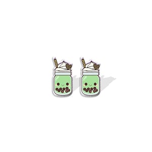 Op de afbeelding: Een paar groene en witte cartoon bubble tea oorbellen met een bruine rietje en een hartje bovenop.