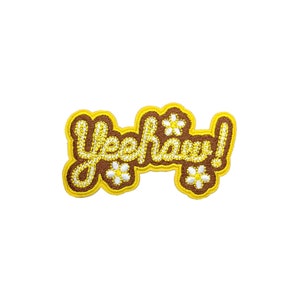 以下が含まれることがあります： 黄色のステッチで「Yeehaw!」という文字が書かれた茶色のフェルトパッチ。パッチは白いデイジーの花で飾られています。