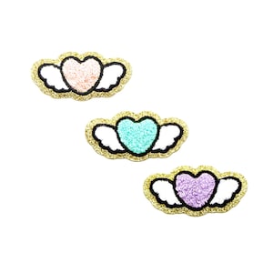 Könnte beinhalten: Drei herzförmige Aufnäher mit Flügeln. Jedes Herz hat eine andere Farbe: rosa, türkis und lila. Die Flügel sind weiß und die Ränder sind mit schwarzem und goldenem Glitzer umrandet.