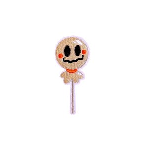 May include: Embroidered patch featuring a smiling, ghost-like lollipop design. The lollipop has a tan face with black eyes and mouth, orange cheeks and collar, and a light brown stick. Perfect for Halloween or spooky-themed crafts.