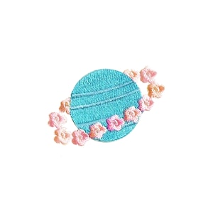 May include: A blue embroidered planet with pink flowers around it. The planet has a ring around it.