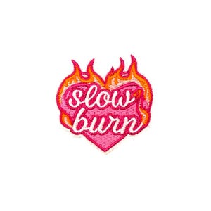 May include: Embroidered patch featuring a pink heart with the words "slow burn" in cursive. The heart is surrounded by orange and yellow flames, creating a fiery effect. The patch is designed for clothing or accessories.
