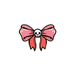 May include: A pink bow-shaped patch with a skull in the center. The bow has a glossy appearance and is outlined in black. The patch is likely for clothing or accessories.