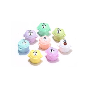 Może przedstawiać: Osiem małych ceramicznych doniczek w pastelowych kolorach z pyszczkami niedźwiedzi i pokrywkami. Doniczki mają uchwyt w kształcie pętli i są ułożone w kółko na białym tle.