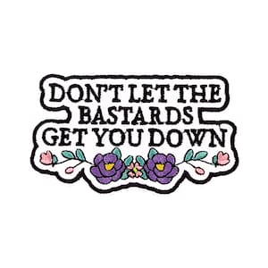 Può includere: Toppa ricamata con la scritta "DON'T LET THE BASTARDS GET YOU DOWN" in nero, bordata di bianco. Presenta accenti floreali viola e rosa su sfondo bianco.