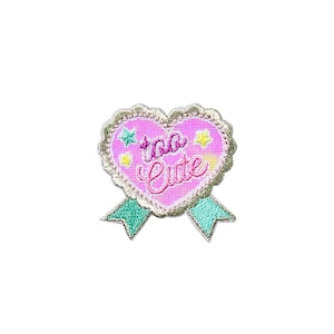 Peut inclure: Patch brodé en forme de cœur rose avec le texte "too cute" et deux étoiles jaunes. Le patch est doté d'un ruban vert attaché en bas.