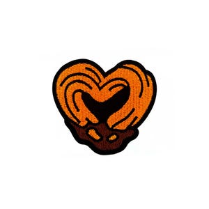 Puede incluir: Parche bordado con forma de pretzel en forma de corazón. El pretzel es naranja con detalles marrón oscuro y una base marrón oscuro similar al chocolate. El parche está sobre un fondo blanco.