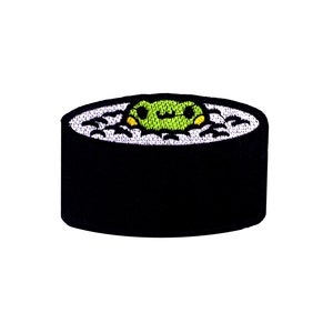 May include: An embroidered patch of a sushi roll with a green seaweed filling and white rice. The sushi roll is black and white.