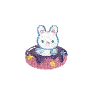 以下が含まれることがあります： ピンクとパープルのドーナツの上に座っている白い刺繍のウサギ。ドーナツには黄色の星が散りばめられています。