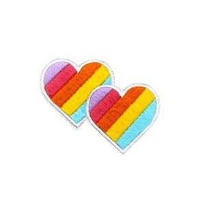 May include: Two embroidered rainbow heart patches. The hearts are outlined in white and filled with stripes of purple, red, orange, yellow, and blue.