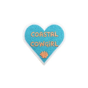 May include: A blue heart-shaped patch with white stitching and the text "COASTAL COWGIRL" in orange. A small orange seashell is in the center of the heart.