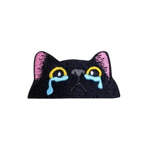 May include: Embroidered patch of a black cat face with yellow eyes and pink ears, shedding blue tears. This patch is designed for application to fabric, ideal for customising clothing or accessories.