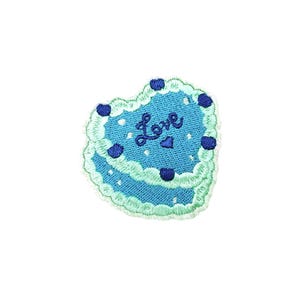Puede incluir: Un parche bordado en forma de corazón azul con glaseado blanco y chispas azules. La palabra "Love" está bordada en azul en el corazón.
