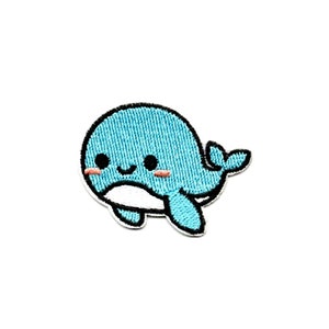 Puede incluir: Un parche bordado de ballena azul y blanco con una cara sonriente y mejillas rosadas. La ballena es de estilo de dibujos animados y tiene una pequeña aleta de cola.