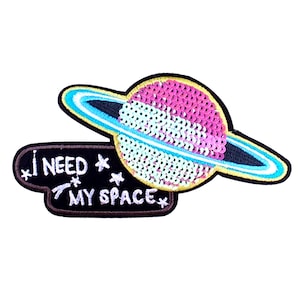 Puede incluir: Un parche bordado en blanco y negro con un planeta de lentejuelas rosa y blanco con un anillo azul y amarillo. El texto "I NEED MY SPACE" está bordado en blanco en el parche.