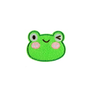 Könnte beinhalten: Ein grüner, gestickter Aufnäher in Form eines Froschgesichts. Der Frosch hat ein zwinkerndes Auge, rosa Wangen und einen lächelnden Mund. Der Aufnäher ist mit dunklerem Grün umrandet.