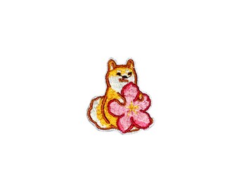 May include: Embroidered patch featuring a cartoon Shiba Inu dog holding a pink flower. The dog has a brown and white fur pattern and a serious expression.