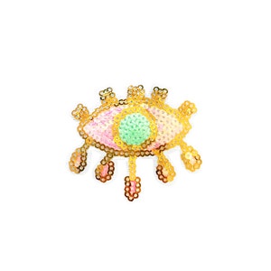 May include: An eye-shaped patch, embellished with gold sequins, a pink and green centre, and dangling gold sequin accents. The patch is set against a white background. This decorative patch is suitable for adding a unique touch to clothing or accessories.