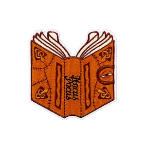 May include: An embroidered patch of a brown book with the words "Hocus Focus" on the spine. The book has a stitched leather cover with a decorative eye and other details.