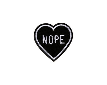 Peut inclure: Patch en forme de cœur noir avec des coutures blanches et le mot "NOPE" en lettres blanches.