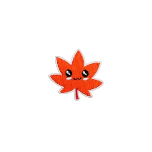 May include: An embroidered patch featuring a cartoon maple leaf with a smiling face. The leaf is orange with a white border and a red stem. The eyes are black and the mouth is a small curved line.