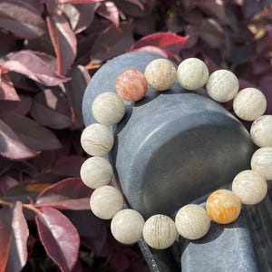 May include: A beaded bracelet featuring round, light-colored beads with subtle patterns and variations in color. The bracelet includes beads in shades of cream, beige, and a few with orange and peach tones. The bracelet is displayed against a dark gray background.