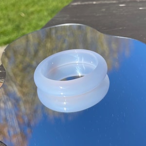 May include: A translucent, light blue ring with a smooth, rounded design. The ring is resting on a reflective surface, creating a mirrored effect. The ring's color and shape suggest it is made of a smooth material, possibly glass or resin.