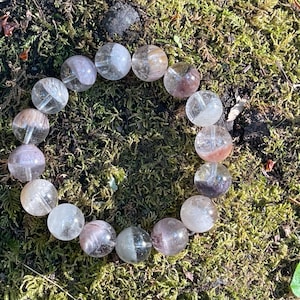 May include: A clear quartz bead bracelet with a variety of colors, including pink, white, and brown. The beads are round and strung together, resting on a bed of green moss.