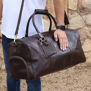 May include: A brown leather duffel bag with a shoulder strap and top handles. The bag has a small pocket on the front.