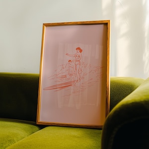 May include: Framed art print featuring a retro illustration of two people surfing. The artwork is rendered in a coral color on a pale pink background. The frame is a warm, natural wood tone, adding a touch of vintage charm to the piece.