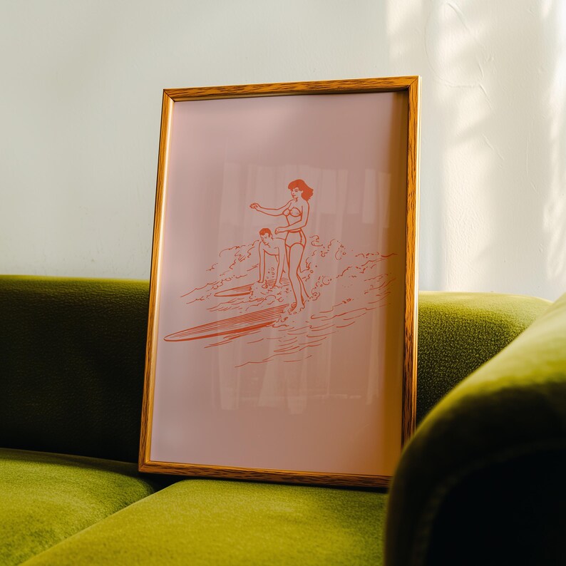 May include: Framed art print featuring a retro illustration of two people surfing. The artwork is rendered in a coral color on a pale pink background. The frame is a warm, natural wood tone, adding a touch of vintage charm to the piece.