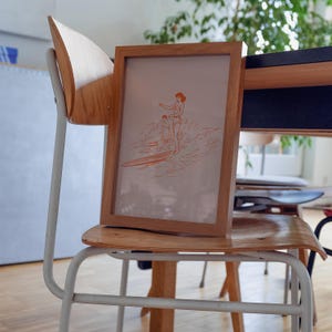 May include: A framed print featuring an orange line drawing of two people surfing. The artwork is in a light brown wooden frame. The frame is resting on a light wood chair with a white metal frame. The background includes a table and a houseplant.