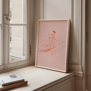 May include: Framed art print featuring a pink background with an orange line drawing of two people surfing. The artwork is in a light wooden frame and is displayed on a light-colored surface near a window.