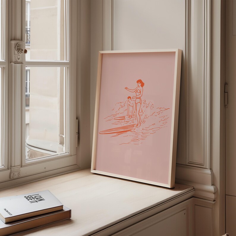 May include: Framed art print featuring a pink background with an orange line drawing of two people surfing. The artwork is in a light wooden frame and is displayed on a light-colored surface near a window.
