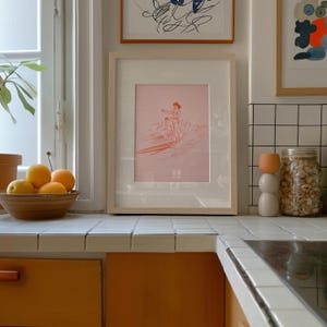 May include: A framed pink print of a surfer on a surfboard, with a light wood frame. The print is displayed on a white tiled countertop in a kitchen setting. A bowl of oranges and a decorative jar are also visible.
