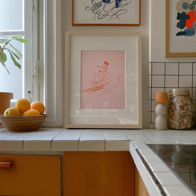 May include: A framed pink print of a surfer on a surfboard, with a light wood frame. The print is displayed on a white tiled countertop in a kitchen setting. A bowl of oranges and a decorative jar are also visible.
