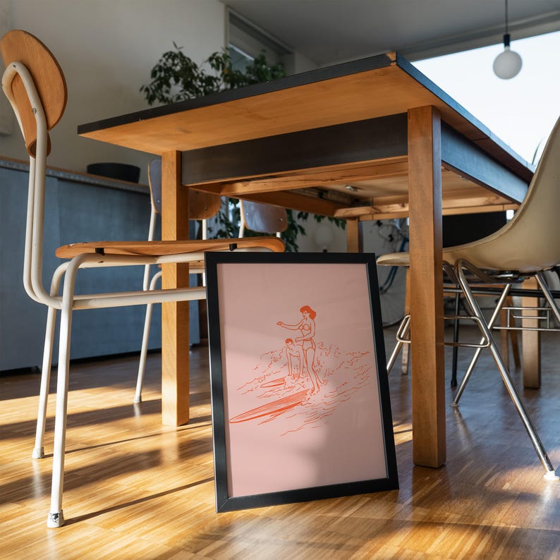 May include: Framed art print featuring a line drawing of two people surfing. The artwork is in shades of orange and is set against a pink background. The black frame is leaning against a wooden floor, with a table and chairs in the background.