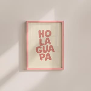 Op de afbeelding: Een ingelijste print met de woorden "HOLA GUAPA PA" in een koraalkleurig lettertype. De print staat tegen een crèmekleurige achtergrond en is ingelijst in een lichtroze lijst. Het kunstwerk wordt op een lichtroze muur getoond.