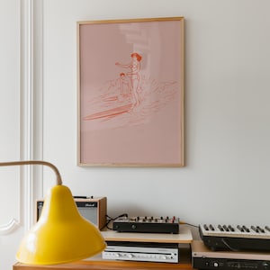 May include: A framed print featuring a coral-colored illustration of two people surfing. The artwork is set against a pale pink background and framed in natural wood. The print hangs on a white wall, above a stereo system and a yellow lamp.
