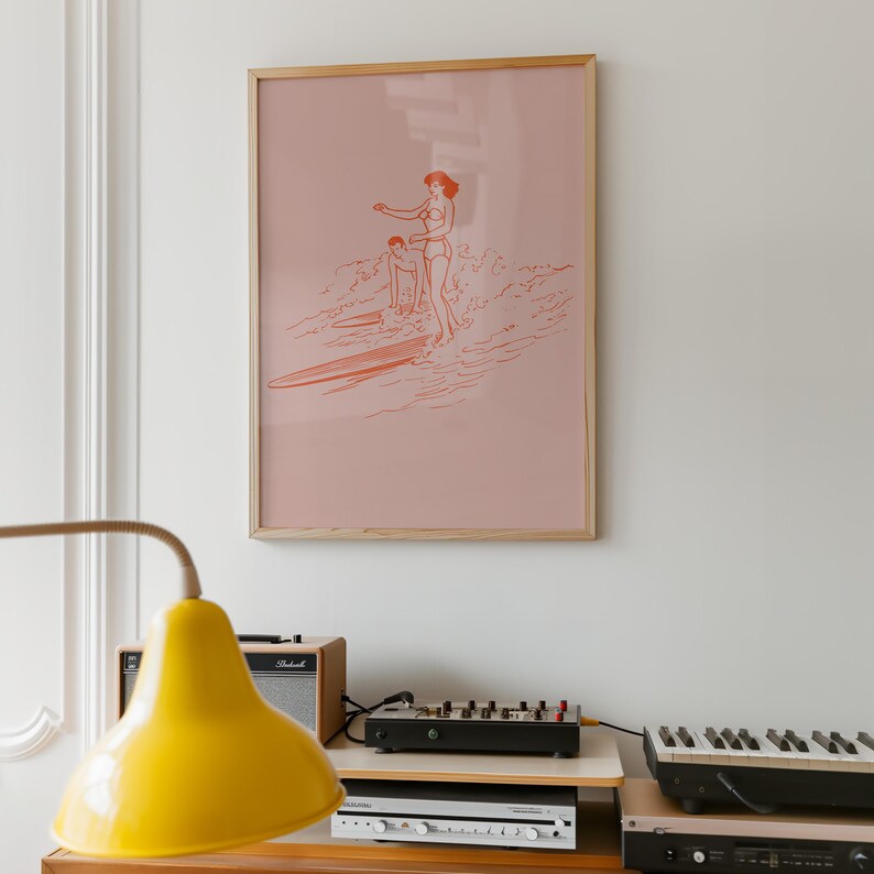 May include: A framed print featuring a coral-colored illustration of two people surfing. The artwork is set against a pale pink background and framed in natural wood. The print hangs on a white wall, above a stereo system and a yellow lamp.