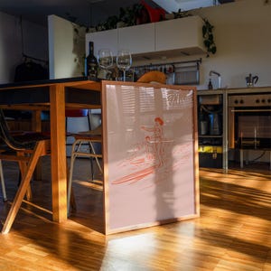 May include: A framed print featuring a red and white illustration of two people surfing. The artwork is set against a pink background and framed in light wood. The print is displayed in a room with a wooden table, chairs, and kitchen appliances.