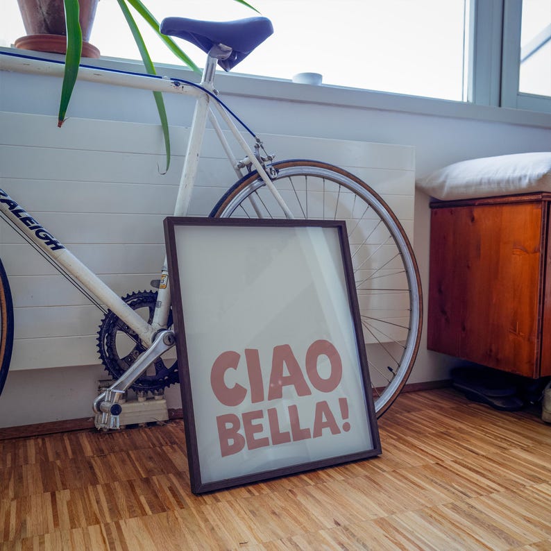 May include: A framed print with the text "CIAO BELLA!" in a pinkish-red font. The frame is dark brown. A white bicycle with a blue seat and brown tyres is behind the frame. The image is on a wooden floor.
