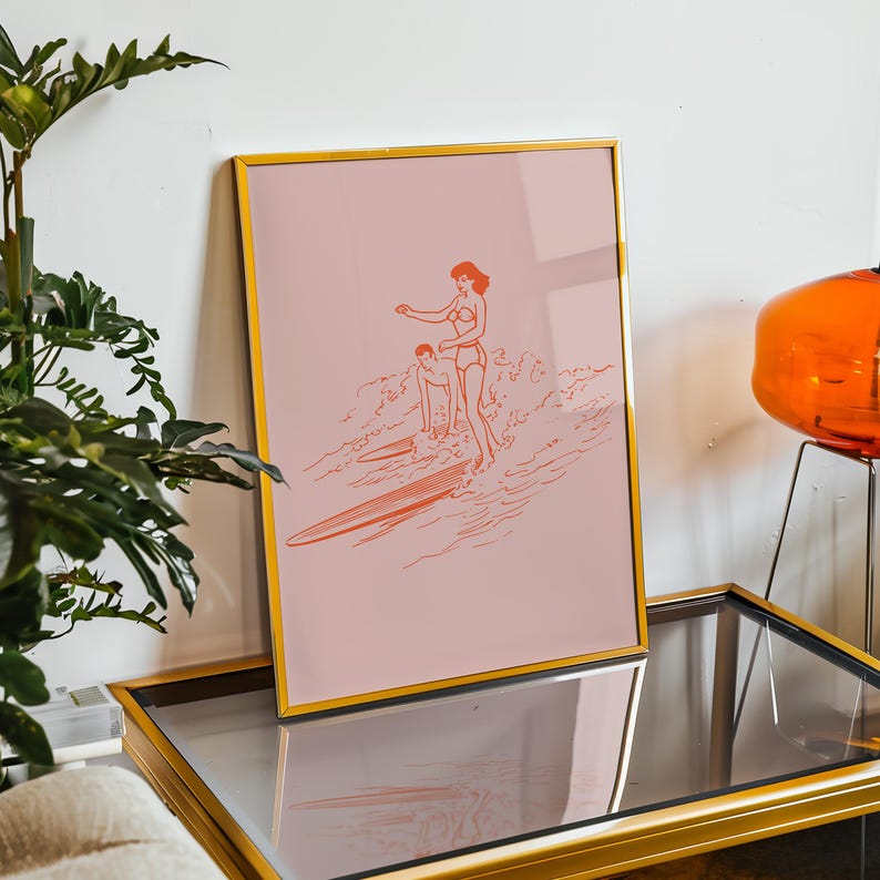 May include: A framed art print featuring a coral-colored illustration of two people surfing on a pink background. The artwork is in a gold frame and depicts a woman and a person on a surfboard in the ocean.