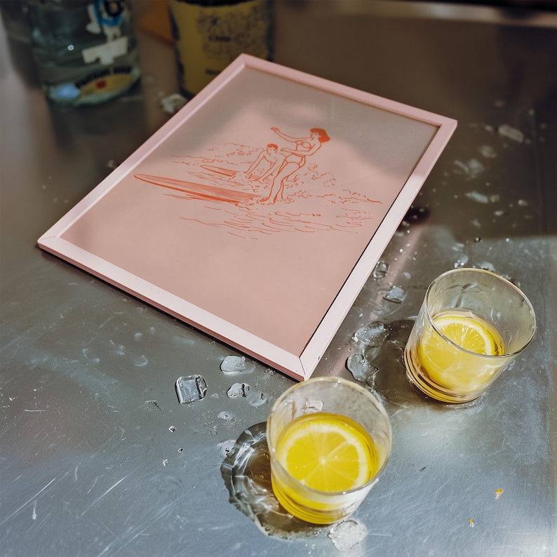 May include: A pink framed print featuring a surfer illustration in orange. Two clear glasses with lemon slices sit next to the frame on a metal surface. Ice cubes and water droplets are scattered around the glasses.