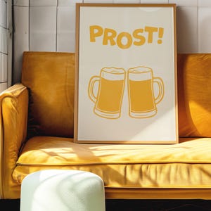 May include: A framed print with the word "PROST!" in yellow letters and two yellow beer mugs clinking together. The print is on a white background and is framed in a light wood frame.