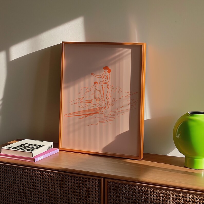 May include: Orange framed art print featuring a minimalist illustration of two people surfing. The artwork depicts figures in swimwear on surfboards in the ocean. The print is displayed on a wooden cabinet with books and a green vase.