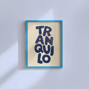 May include: A framed art print featuring the word "TRANQUILO" in dark blue, bold lettering. The print has a light beige background and is set within a bright blue frame. The artwork is displayed on a white wall.