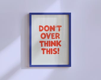 May include: A framed print with the words "DON'T OVER THINK THIS!" in bold red letters. The text is centered on a white background, framed by a blue border. The print is hanging on a light purple wall.